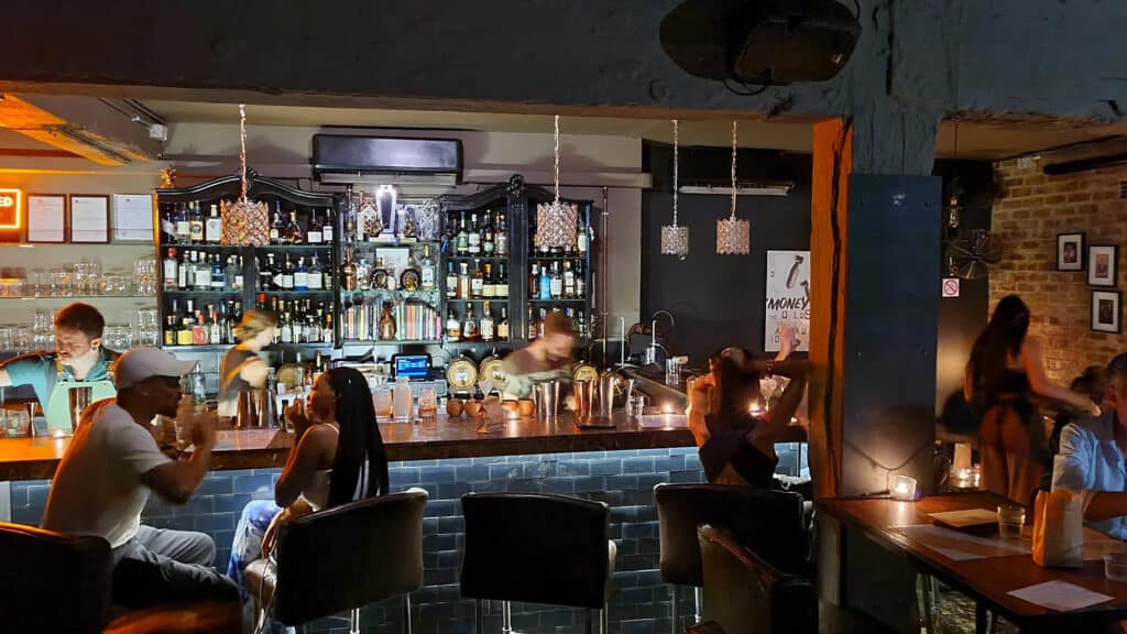 A view of Sin + Tax bar in Rosebank, featuring a stylish bar counter, bar attendants preparing drinks, and patrons seated around the bar enjoying the vibrant atmosphere.